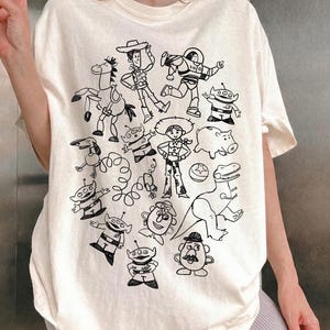 Camiseta unisex de Disney Pixar Toy Story Doodles Comfort Colors, camiseta de dibujos animados de Woody y Buzz Lightyear, camiseta a juego para regalar en un viaje familiar a Disneyland. Natural/ Ivory