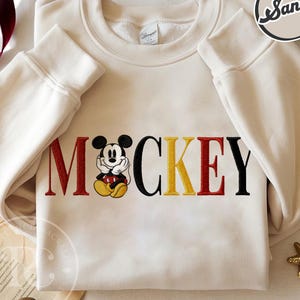 以下が含まれることがあります： 赤、黒、黄色で「MICKEY」の文字が刺繍されたクリーム色のスウェットシャツ。漫画のキャラクターが特徴です。長袖トップスはクルーネックとリブ編みの袖口が付いています。
