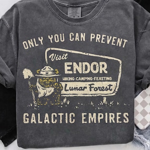 May include: Dark grey sweatshirt with the text "ONLY YOU CAN PREVENT" above a sign that reads "Visit ENDOR Hiking-Camping-Feasting Lunar Forest" and "GALACTIC EMPIRES" below. The sign features a cartoon character with a shovel.