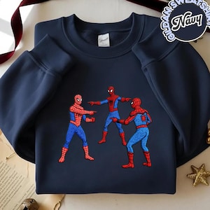 Puede incluir: Sudadera azul marino con un dise&ntilde;o bordado de tres personajes de Spider-Man se&ntilde;al&aacute;ndose. La sudadera tiene mangas largas y cuello redondo. El texto "Gildan Sweatshirt Navy" est&aacute; impreso en la esquina superior derecha.