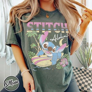Puede incluir: Camiseta verde oscuro con un personaje de dibujos animados de Stitch surfeando. La camiseta tiene la palabra "STITCH" en rosa y la palabra "Aloha" en blanco. Tambi&eacute;n se ven palmeras y una luna creciente.