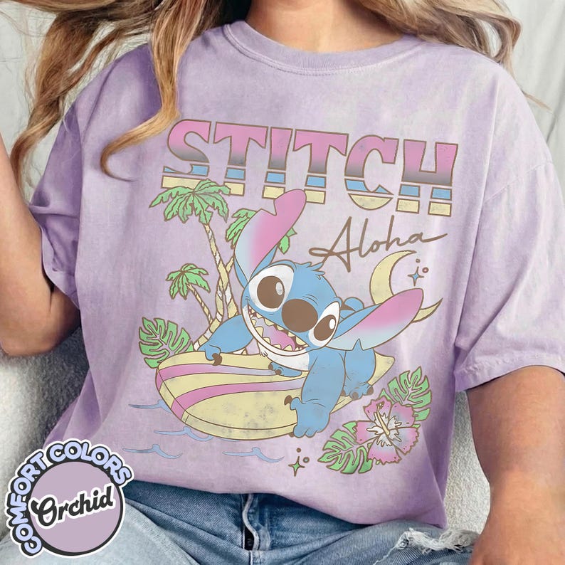 Puede incluir: Camiseta color orqu&iacute;dea con un gr&aacute;fico de Stitch surfeando. El dise&ntilde;o incluye la palabra "STITCH" en una fuente retro, la palabra "Aloha", palmeras, una luna creciente y una flor de hibisco. La camiseta tiene una etiqueta Comfort Colors.