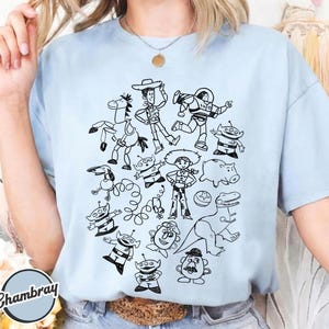 Puede incluir: Camiseta azul claro con un garabato en blanco y negro de personajes de dibujos animados. El dise&ntilde;o incluye un vaquero, un guardabosques espacial, extraterrestres y otras figuras. La camiseta tiene mangas cortas y cuello redondo.