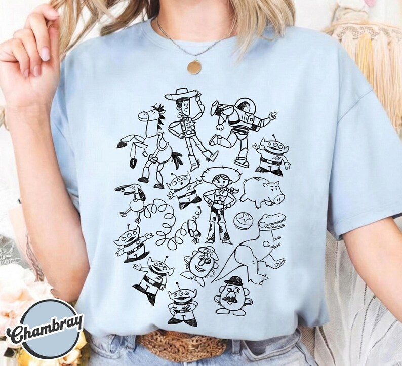 Puede incluir: Camiseta azul claro con un garabato en blanco y negro de personajes de dibujos animados. El dise&ntilde;o incluye un vaquero, un guardabosques espacial, extraterrestres y otras figuras. La camiseta tiene mangas cortas y cuello redondo.