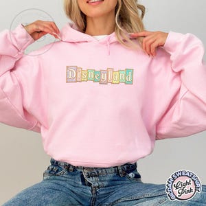 May include: A light pink hooded sweatshirt with the word "Disneyland" embroidered across the chest in pastel colors. The sweatshirt has a drawstring hood and long sleeves. The text is in a block letter style.