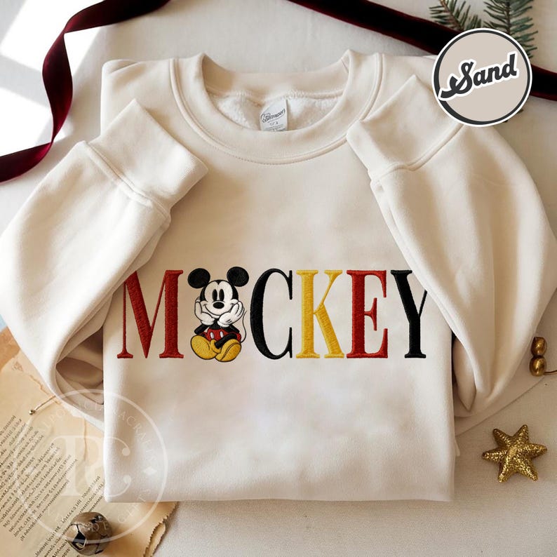 以下が含まれることがあります： 赤、黒、黄色で「MICKEY」の文字が刺繍されたクリーム色のスウェットシャツ。漫画のキャラクターが特徴です。長袖トップスはクルーネックとリブ編みの袖口が付いています。