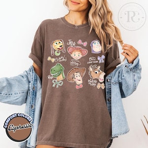 K&ouml;nnte beinhalten: Espressofarbenes T-Shirt mit Cartoon-Illustrationen von Charakteren aus dem Film Toy Story. Das Shirt enth&auml;lt Bilder von Woody, Buzz Lightyear, Jessie, Rex und anderen. Auf dem Shirt stehen auch die Worte "Comfort Colors" und "Espresso".
