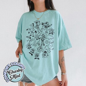 Puede incluir: Camiseta azul claro con una ilustraci&oacute;n en blanco y negro de varios personajes de dibujos animados. La camiseta tiene mangas cortas y un corte holgado. El texto "COMFORT COLORS" y "Charky Mint" est&aacute; impreso en la parte inferior izquierda.