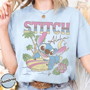 Puede incluir: Camiseta azul claro con un dibujo de Stitch surfeando en una tabla de surf. La camiseta tiene el texto "STITCH Aloha" en una fuente retro, con palmeras, flores y una luna creciente.