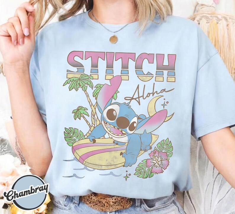 Puede incluir: Camiseta azul claro con un dibujo de Stitch surfeando en una tabla de surf. La camiseta tiene el texto "STITCH Aloha" en una fuente retro, con palmeras, flores y una luna creciente.
