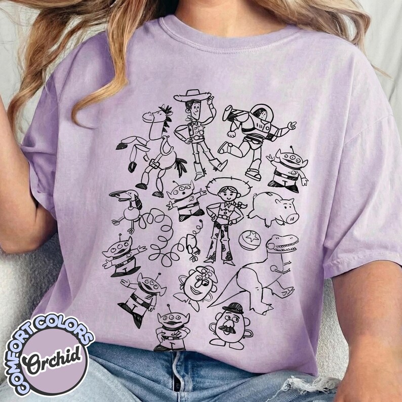 Puede incluir: Camiseta color orqu&iacute;dea con un dibujo lineal negro de varios personajes de dibujos animados. El dise&ntilde;o incluye un vaquero, un astronauta, extraterrestres y otras figuras. La camiseta tiene una etiqueta Comfort Colors.