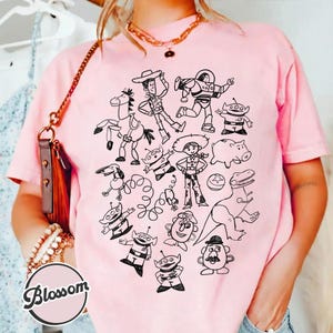 Puede incluir: Camiseta rosa claro con un dibujo lineal negro de personajes de Toy Story. El dise&ntilde;o incluye a Woody, Buzz Lightyear y otros personajes. La camiseta tiene cuello redondo y mangas cortas.