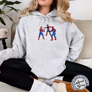 Puede incluir: Sudadera con capucha gris claro con un dise&ntilde;o bordado de tres figuras de Spider-Man se&ntilde;al&aacute;ndose. La sudadera est&aacute; hecha de un material suave y tiene una capucha con cord&oacute;n. La etiqueta Gildan Sport Grey es visible en la parte inferior derecha.