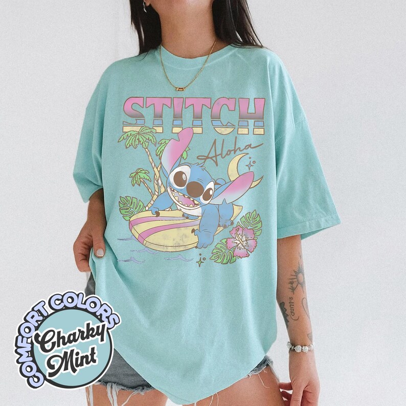 Puede incluir: Camiseta azul claro con un dise&ntilde;o de Stitch surfeando y el texto "STITCH Aloha". El dise&ntilde;o incluye palmeras, una luna creciente y una flor rosa. La camiseta tiene mangas cortas y cuello redondo.