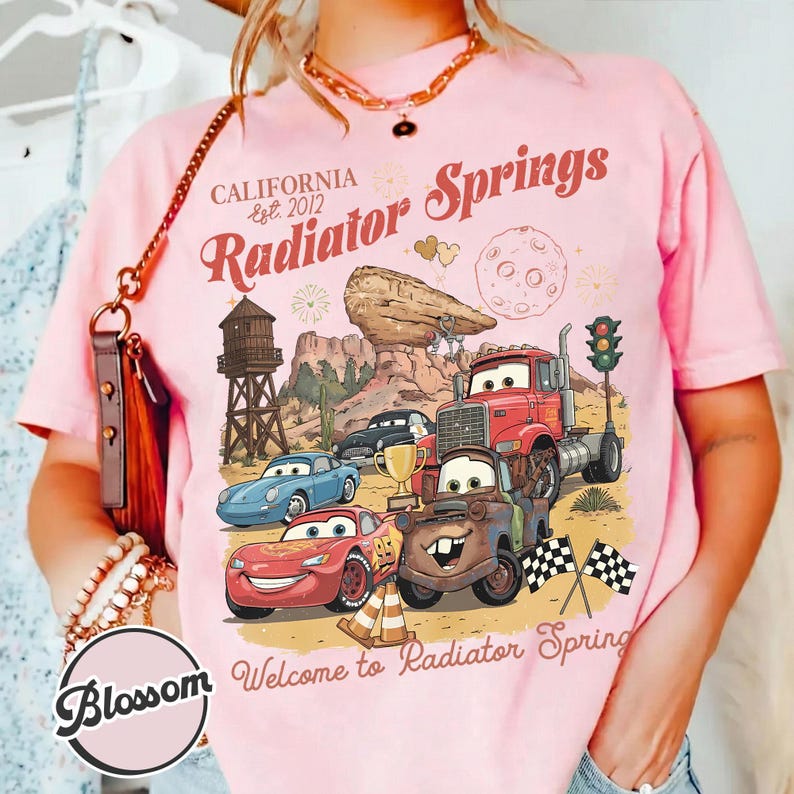 Pode incluir: T-shirt rosa com um design gr&aacute;fico com personagens do filme Carros, com o texto "California Est. 2012 Radiator Springs". O design inclui carros de desenho animado, uma paisagem des&eacute;rtica e a frase "Welcome to Radiator Spring".