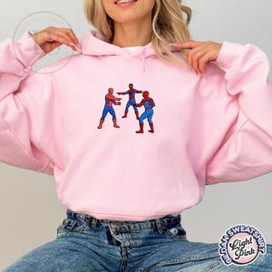 Puede incluir: Sudadera con capucha rosa claro con un dise&ntilde;o bordado de tres figuras de Spider-Man. Las figuras visten trajes rojos y azules. La sudadera tiene una capucha con cord&oacute;n y mangas largas. El dise&ntilde;o est&aacute; centrado en el pecho.