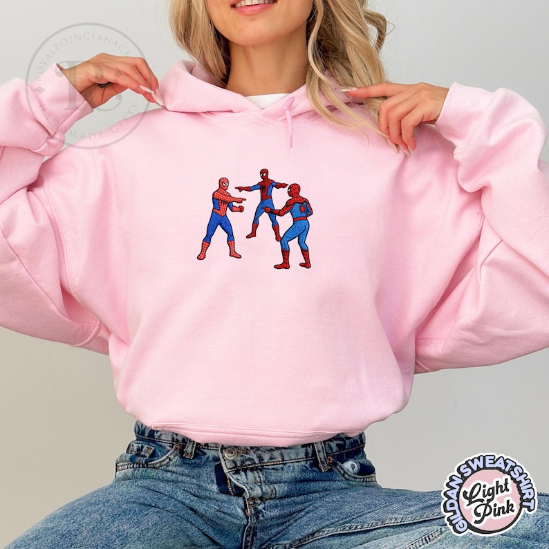 Puede incluir: Sudadera con capucha rosa claro con un dise&ntilde;o bordado de tres figuras de Spider-Man. Las figuras visten trajes rojos y azules. La sudadera tiene una capucha con cord&oacute;n y mangas largas. El dise&ntilde;o est&aacute; centrado en el pecho.