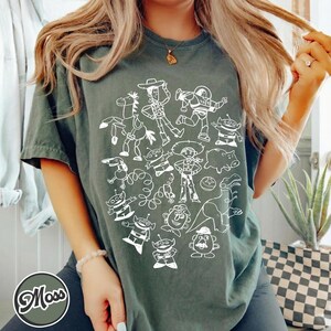 Puede incluir: Camiseta verde oscuro con una ilustraci&oacute;n en blanco de personajes de una popular pel&iacute;cula de animaci&oacute;n. El dise&ntilde;o incluye un vaquero, un guardabosques espacial y otras figuras reconocibles. La camiseta tiene mangas cortas y cuello redondo.