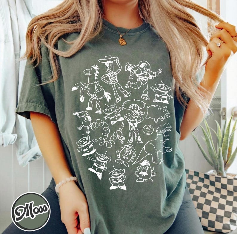 Puede incluir: Camiseta verde oscuro con una ilustraci&oacute;n en blanco de personajes de una popular pel&iacute;cula de animaci&oacute;n. El dise&ntilde;o incluye un vaquero, un guardabosques espacial y otras figuras reconocibles. La camiseta tiene mangas cortas y cuello redondo.
