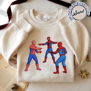 Puede incluir: Sudadera color crema con un dise&ntilde;o bordado de tres figuras de Spider-Man se&ntilde;al&aacute;ndose entre s&iacute;. Las figuras de Spider-Man visten sus trajes cl&aacute;sicos rojo y azul. La sudadera tiene mangas largas y cuello redondo.