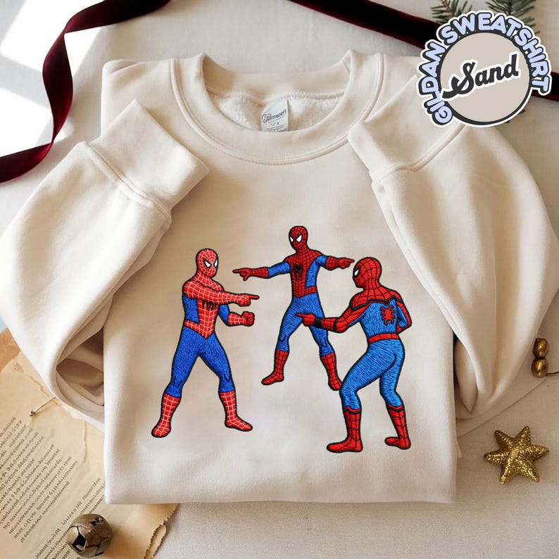 Puede incluir: Sudadera color crema con un dise&ntilde;o bordado de tres figuras de Spider-Man se&ntilde;al&aacute;ndose entre s&iacute;. Las figuras de Spider-Man visten sus trajes cl&aacute;sicos rojo y azul. La sudadera tiene mangas largas y cuello redondo.