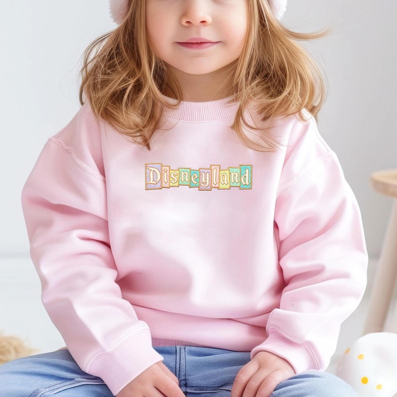 May include: A light pink sweatshirt with the word "Disneyland" embroidered across the chest in pastel colors. The sweatshirt is long-sleeved and appears to be made of a soft material. The design is suitable for children.