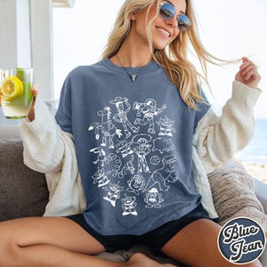 Puede incluir: Una camiseta azul con un dibujo lineal blanco de personajes de dibujos animados. La camiseta tiene un corte holgado y est&aacute; hecha de una tela suave. Las palabras "Blue Jean" est&aacute;n impresas en la esquina inferior derecha de la camiseta.