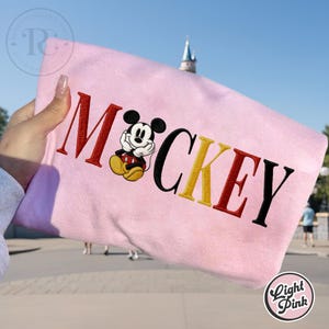 以下が含まれることがあります： 「ミッキー」の文字と漫画のキャラクターが刺繍されたライトピンクのスウェットシャツ。文字は赤、黒、黄色です。ぼやけた背景の前にスウェットシャツが掲げられています。