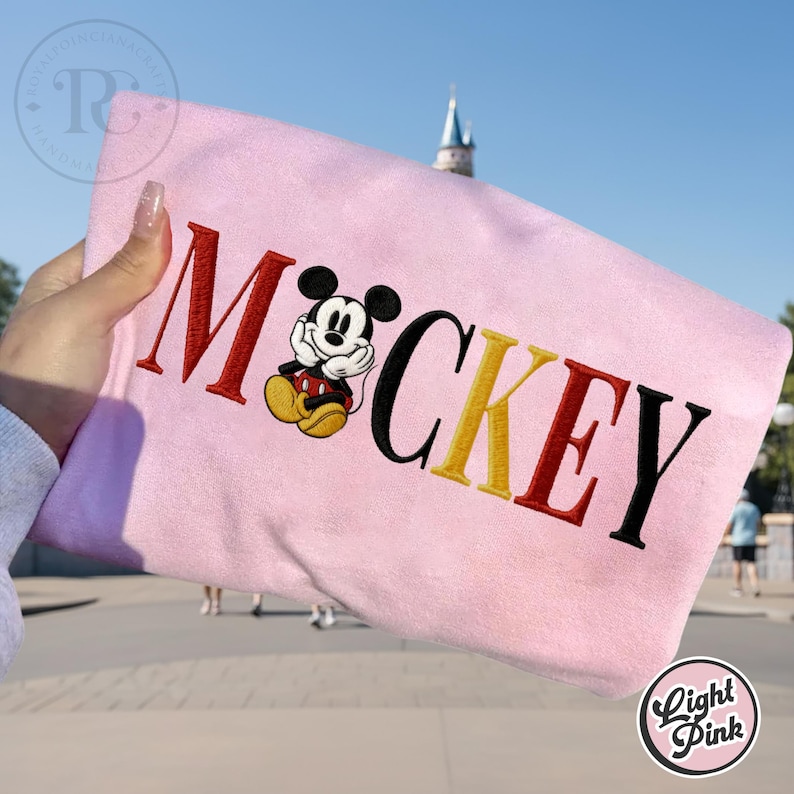 以下が含まれることがあります： 「ミッキー」の文字と漫画のキャラクターが刺繍されたライトピンクのスウェットシャツ。文字は赤、黒、黄色です。ぼやけた背景の前にスウェットシャツが掲げられています。