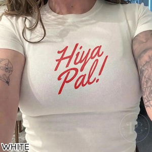 K&ouml;nnte beinhalten: Wei&szlig;es T-Shirt mit dem roten Text "Hiya Pal!". Das Shirt hat einen Rundhalsausschnitt und scheint aus einem weichen, leichten Material zu bestehen. Der Text ist in einer Schreibschrift.