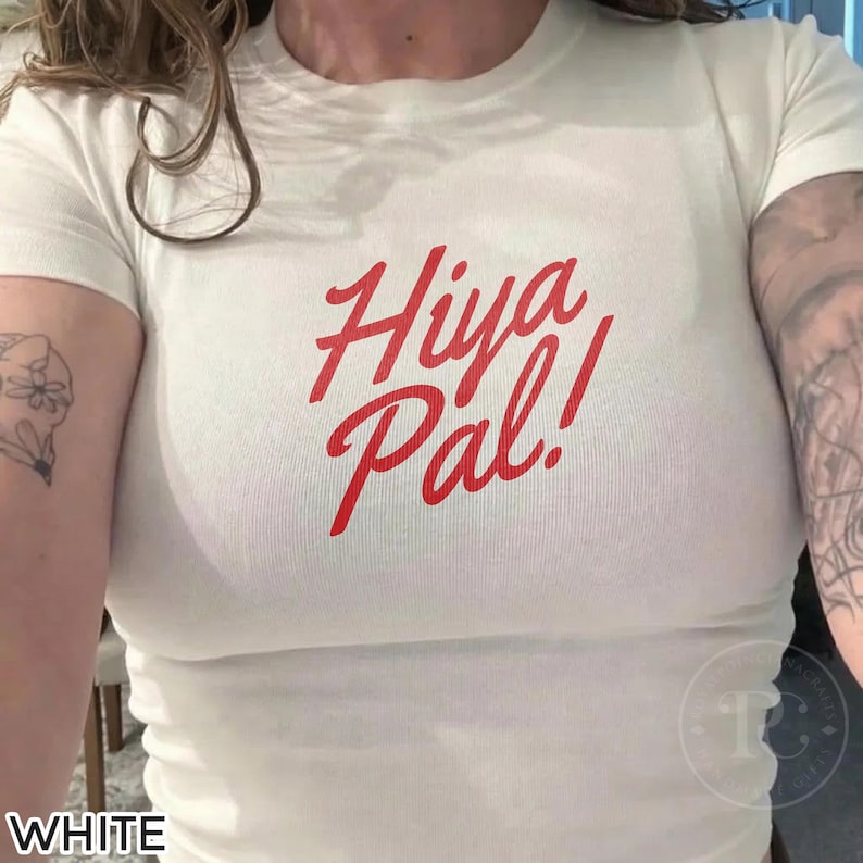 K&ouml;nnte beinhalten: Wei&szlig;es T-Shirt mit dem roten Text "Hiya Pal!". Das Shirt hat einen Rundhalsausschnitt und scheint aus einem weichen, leichten Material zu bestehen. Der Text ist in einer Schreibschrift.