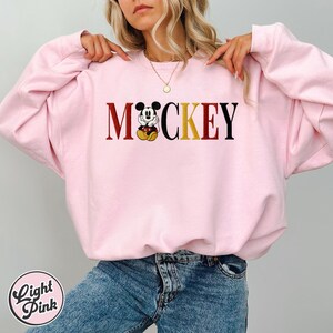 以下が含まれることがあります： スタイリッシュなフォントで「Mickey」という文字がプリントされたライトピンクのスウェットシャツ。「M」と「Y」は濃い赤色、「CKE」は黒色、「O」は黄色で、ミッキーマウスのグラフィックに置き換えられています。「Light Pink」の文字が下部の円の中にあります。