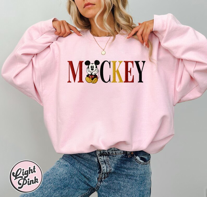 以下が含まれることがあります： スタイリッシュなフォントで「Mickey」という文字がプリントされたライトピンクのスウェットシャツ。「M」と「Y」は濃い赤色、「CKE」は黒色、「O」は黄色で、ミッキーマウスのグラフィックに置き換えられています。「Light Pink」の文字が下部の円の中にあります。