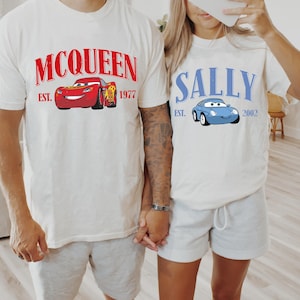 May include: Two white t-shirts with cartoon car graphics. One shirt features a red car with "McQueen" and "Est. 1977" in red text. The other shirt displays a blue car with "Sally" and "Est. 2002" in blue text.