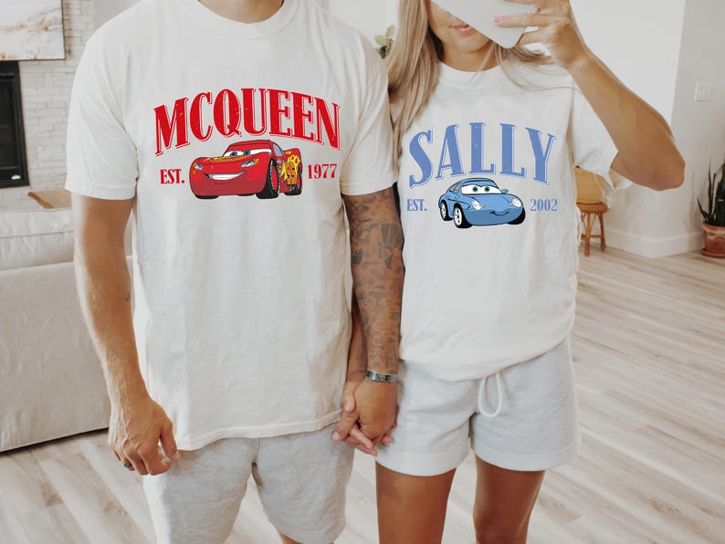 May include: Two white t-shirts with cartoon car graphics. One shirt features a red car with "McQueen" and "Est. 1977" in red text. The other shirt displays a blue car with "Sally" and "Est. 2002" in blue text.