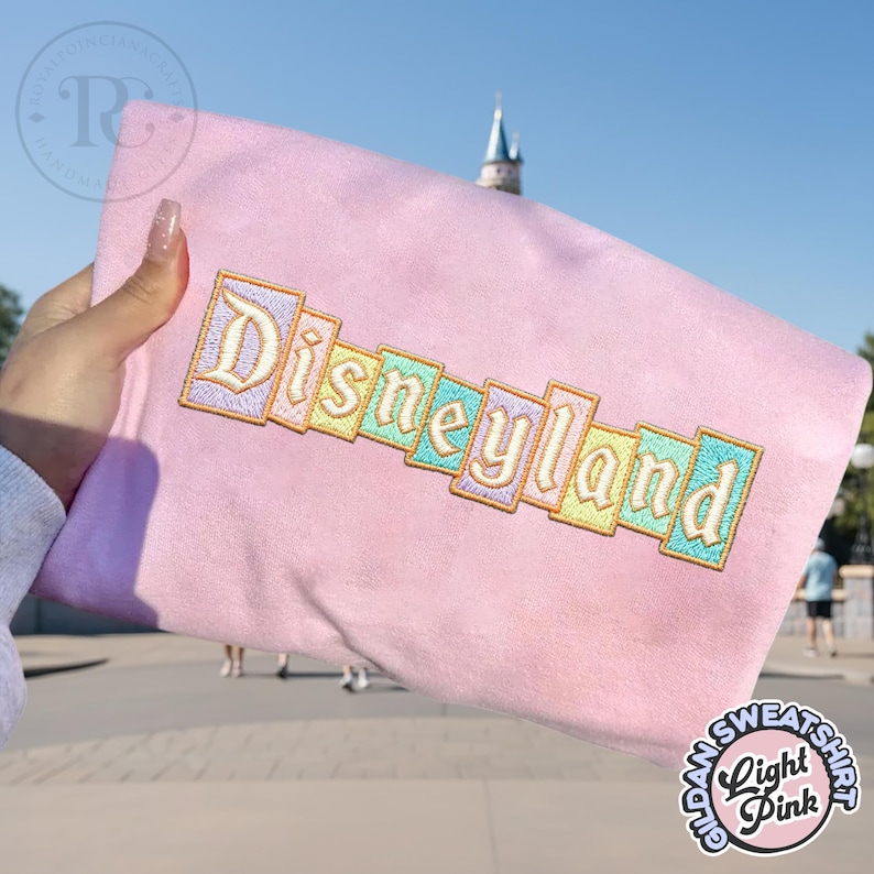 May include: Light pink sweatshirt with the word "Disneyland" embroidered in pastel colors. The text is arranged in a diagonal pattern. The sweatshirt has a soft, plush texture and a round logo that says "Glow Sweatshirt Light Pink".