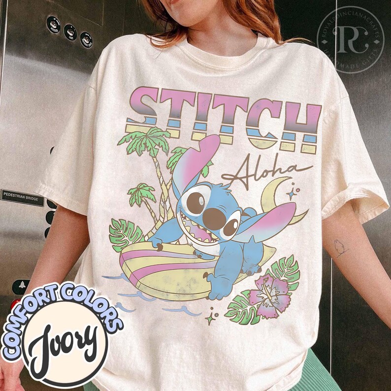 Puede incluir: Camiseta de color marfil con un gr&aacute;fico de Stitch surfeando, con las palabras "STITCH Aloha" en una fuente retro colorida. El dise&ntilde;o incluye palmeras, una luna creciente y flores tropicales. La camiseta es de Comfort Colors.