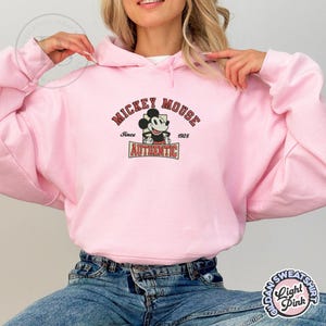 May include: Light pink hooded sweatshirt featuring a Mickey Mouse graphic. The graphic includes the words "Mickey Mouse" above an image of Mickey, with "Since 1928" and "Authentic" below. The sweatshirt is made of a soft material.