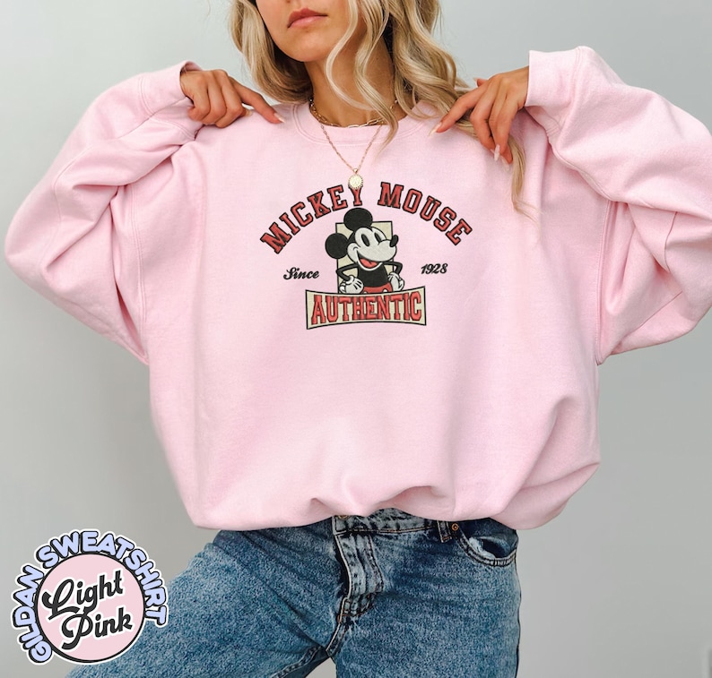 May include: Light pink sweatshirt featuring a Mickey Mouse graphic. The graphic includes the words "Mickey Mouse" and "Authentic" with Mickey Mouse in the center. The sweatshirt also has the words "Since 1928". The sweatshirt is a Gildan brand.