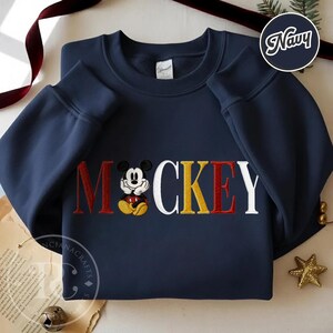 以下が含まれることがあります： 刺繍された「ミッキー」のデザインが施されたネイビーブルーのスウェットシャツ。「ミッキー」の文字は赤、黄色、白で、中央にミッキーマウスがいます。スウェットシャツは長袖で、クルーネックです。