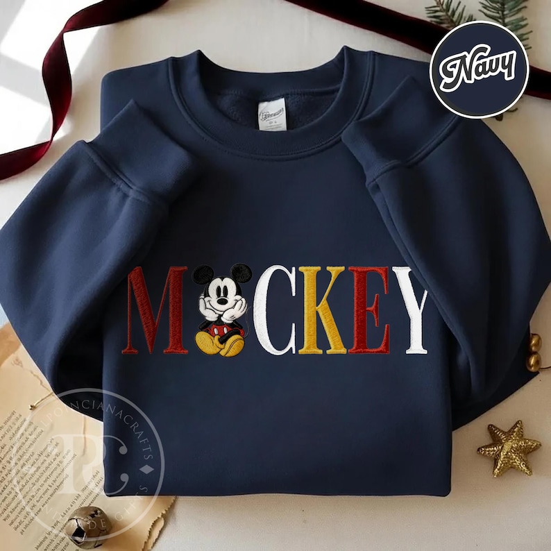 以下が含まれることがあります： 刺繍された「ミッキー」のデザインが施されたネイビーブルーのスウェットシャツ。「ミッキー」の文字は赤、黄色、白で、中央にミッキーマウスがいます。スウェットシャツは長袖で、クルーネックです。