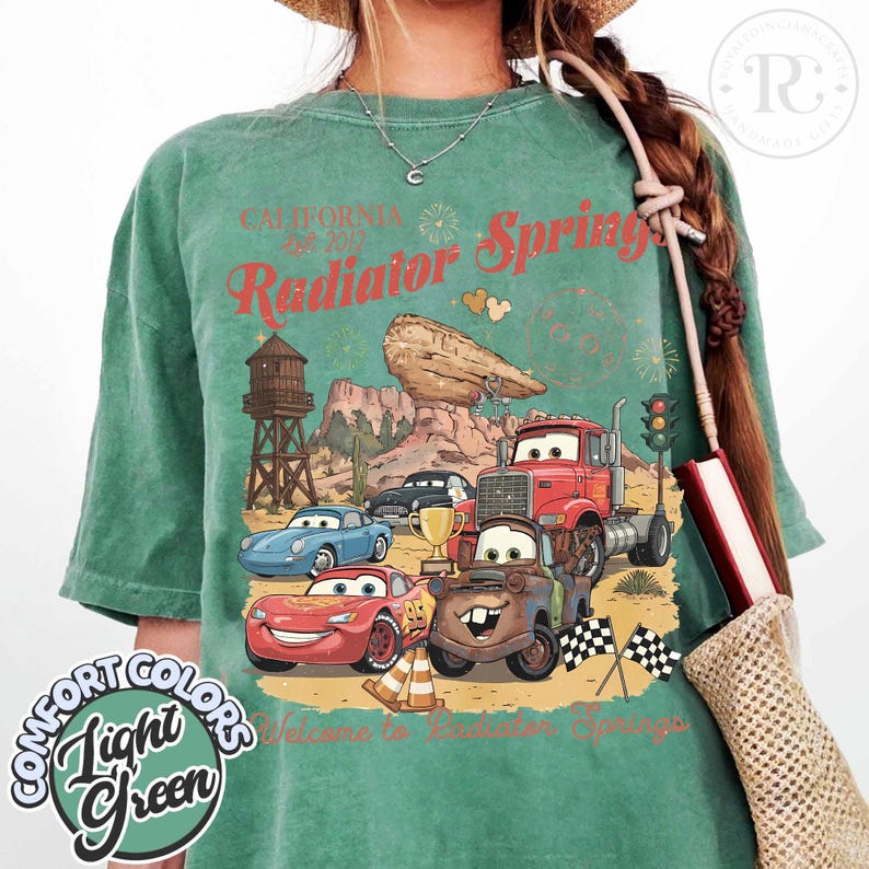 Pode incluir: T-shirt verde claro com um gr&aacute;fico de Radiator Springs do filme Carros. O design inclui carros de desenho animado, uma paisagem des&eacute;rtica e o texto "California Radiator Springs". A camisa tamb&eacute;m tem o texto "Comfort Colors Light Green".