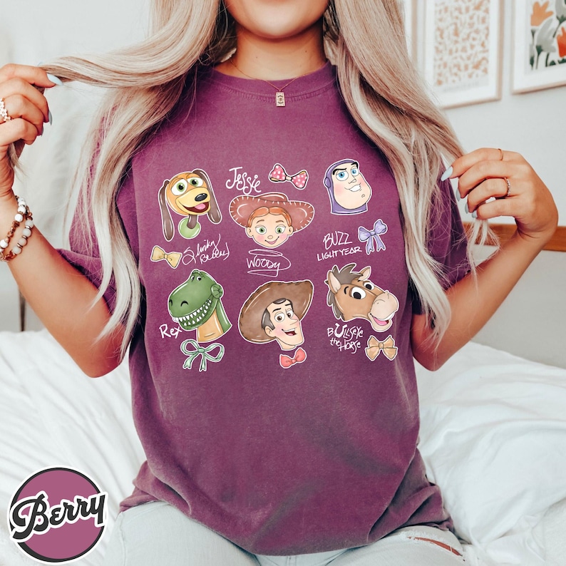 K&ouml;nnte beinhalten: Ein lila T-Shirt mit Illustrationen von Zeichentrickfiguren aus einem beliebten Animationsfilm. Das Shirt zeigt Bilder von Slinky Dog, Jessie, Buzz Lightyear, Rex, Woody und Bullseye the Horse, mit ihren Namen.