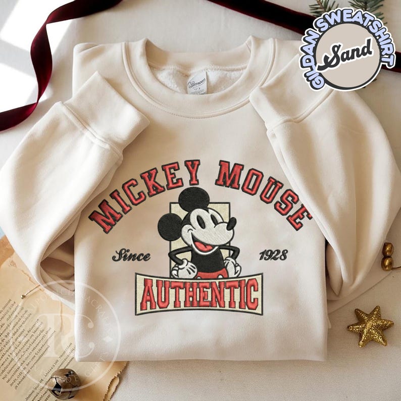 May include: Cream-colored sweatshirt featuring a Mickey Mouse graphic. The design includes the words "Mickey Mouse" in red arched lettering, the character's image, and the word "AUTHENTIC" below. The sweatshirt also has the text "Since 1928".