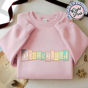 May include: Light pink Gildan sweatshirt with the word "Disneyland" embroidered across the chest in pastel colors. The sweatshirt has long sleeves and a crew neck. A circular graphic reads "Gildan Sweatshirt Light Pink."