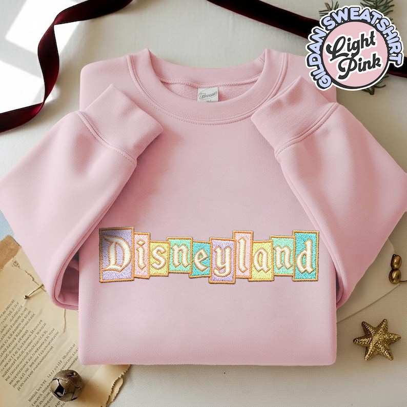 May include: Light pink Gildan sweatshirt with the word "Disneyland" embroidered across the chest in pastel colors. The sweatshirt has long sleeves and a crew neck. A circular graphic reads "Gildan Sweatshirt Light Pink."