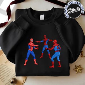 Puede incluir: Sudadera negra de cuello redondo con un gr&aacute;fico de tres personajes de Spider-Man se&ntilde;al&aacute;ndose. Las figuras de Spider-Man visten sus cl&aacute;sicos trajes rojo y azul. La sudadera tiene el texto "Gildan Sweatshirt Black" en un dise&ntilde;o circular.
