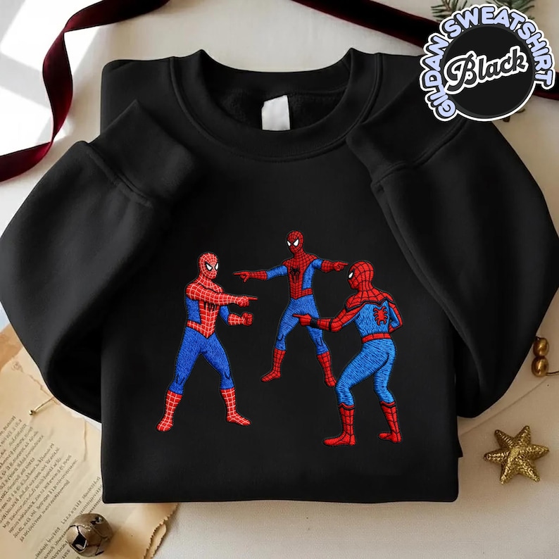 Puede incluir: Sudadera negra de cuello redondo con un gr&aacute;fico de tres personajes de Spider-Man se&ntilde;al&aacute;ndose. Las figuras de Spider-Man visten sus cl&aacute;sicos trajes rojo y azul. La sudadera tiene el texto "Gildan Sweatshirt Black" en un dise&ntilde;o circular.