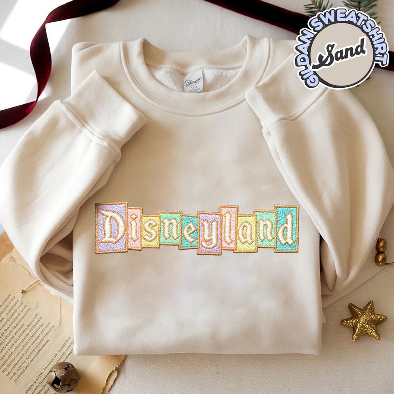 May include: A cream-colored sweatshirt with the word "Disneyland" embroidered across the chest in pastel colors. The sleeves are long and cuffed. The sweatshirt is made of a soft material.