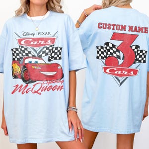 Może przedstawiać: Jasnoniebieskie koszulki z filmu Disney Pixar Cars. Prz&oacute;d koszulki przedstawia Zygzaka McQueena z napisem "Lightning McQueen". Tył koszulki ma napis "CUSTOM NAME" nad dużą czerwoną cyfrą 3 i słowem "Cars".
